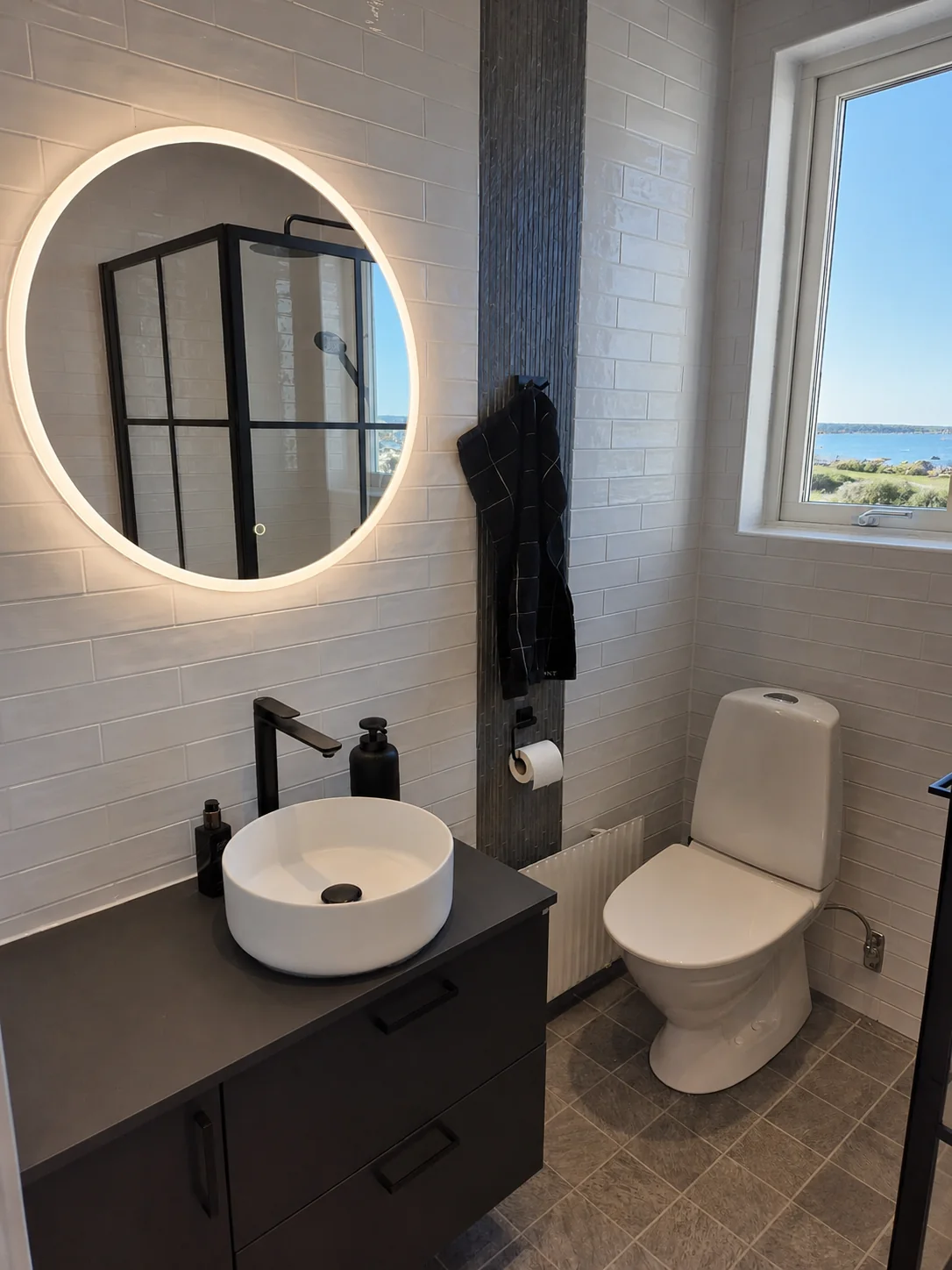 Renoverat badrum med rund LED-spegel och svarta detaljer